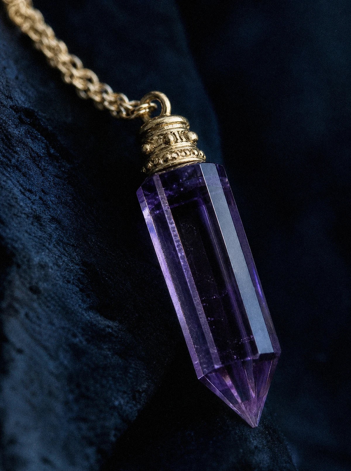 Nahaufnahme eines violetten Kristallanhängers in Form einer sechskantigen Kristallspitze mit goldener Schraubkappe und einem Stück feiner Goldkette, Seitenlicht auf den Kanten, Hintergrund unscharf